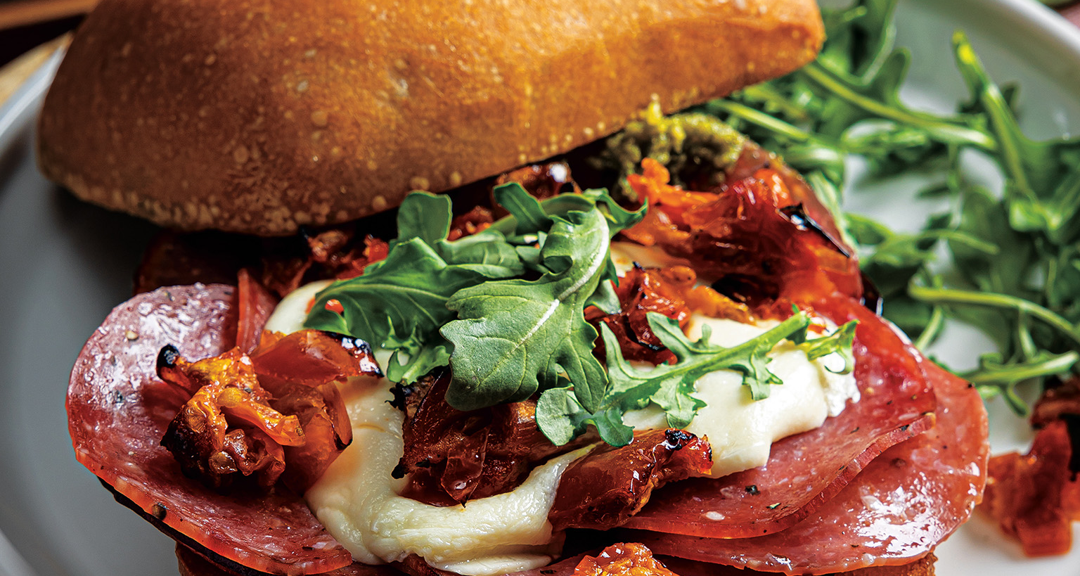  A half-assembled salami and pesto sandwich on a round wooden board with arugula. In the background, a baking sheet holds a second sandwich half with pesto, and another with salami, mozzarella, and sun-dried tomatoes.