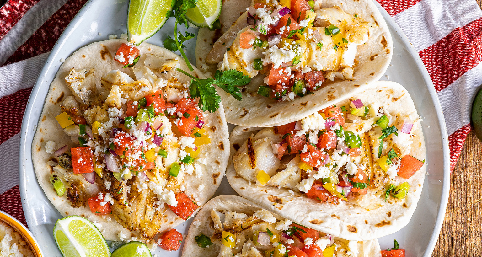 Four vibrant grilled fish street tacos are arranged on a light grey plate, each filled with white fish, colorful watermelon pico de gallo, and crumbled white cheese.