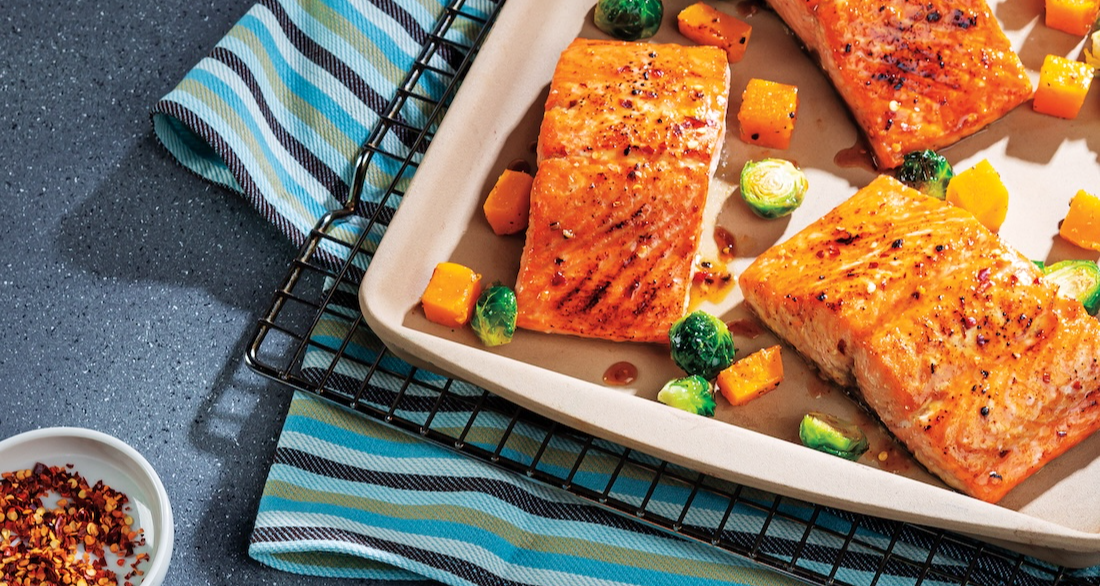 Sheet-pan dinner with honey-mustard glazed salmon and roasted vegetables. A single serving is on a light blue plate with a fork, while the rest of the meal is on a baking sheet in the background.
