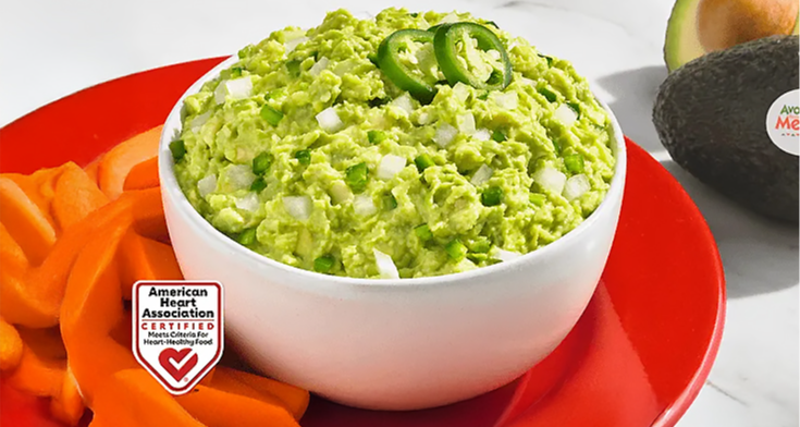  A close-up of a bowl of chunky guacamole topped with jalapeño slices, surrounded by carrot and bell pepper slices on a red plate. An American Heart Association Certified label is on the upper right.