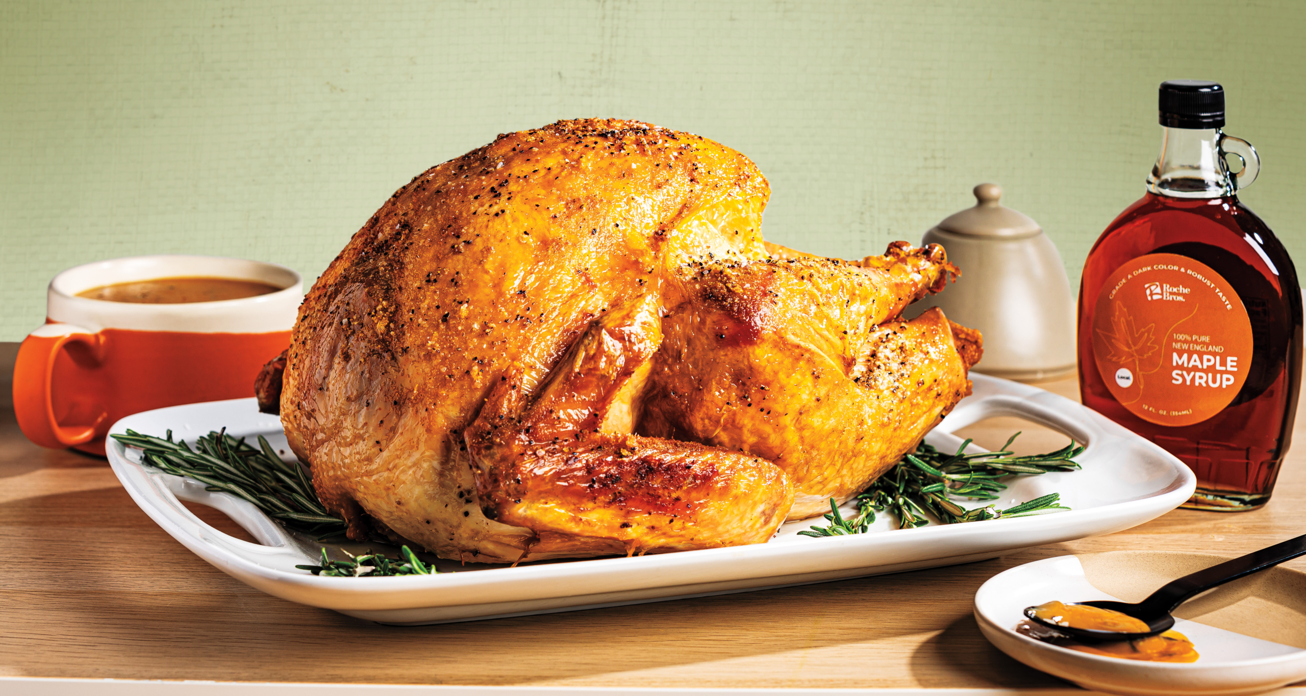 A beautifully roasted turkey with golden-brown skin sits on a white platter garnished with rosemary. To the left is an orange mug of gravy, and to the right is a bottle of maple syrup and a spoon with caramel sauce.