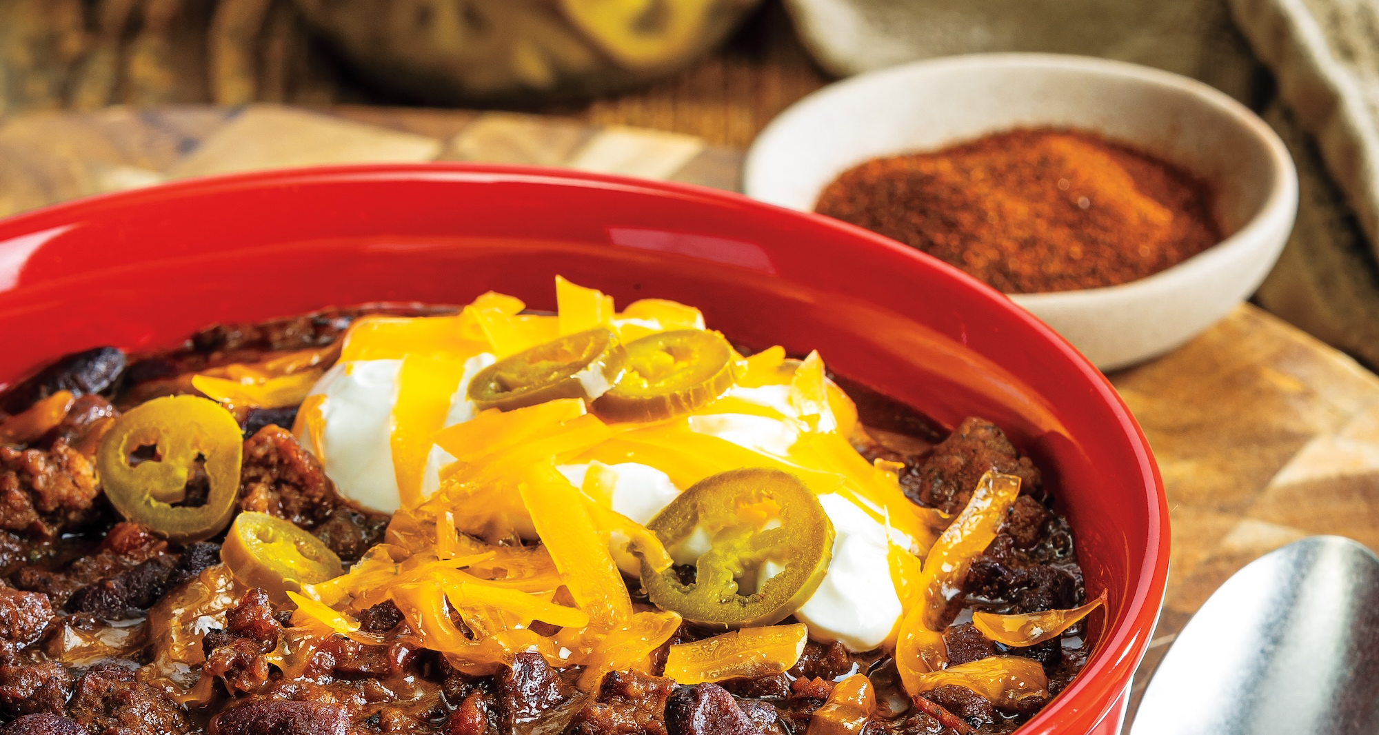 A bright red bowl of beef chili topped with shredded cheese, sour cream, and sliced pickled jalapeños sits on a wooden board next to a spoon. A jar of pickled jalapeños and a small dish of chili powder are in the background.