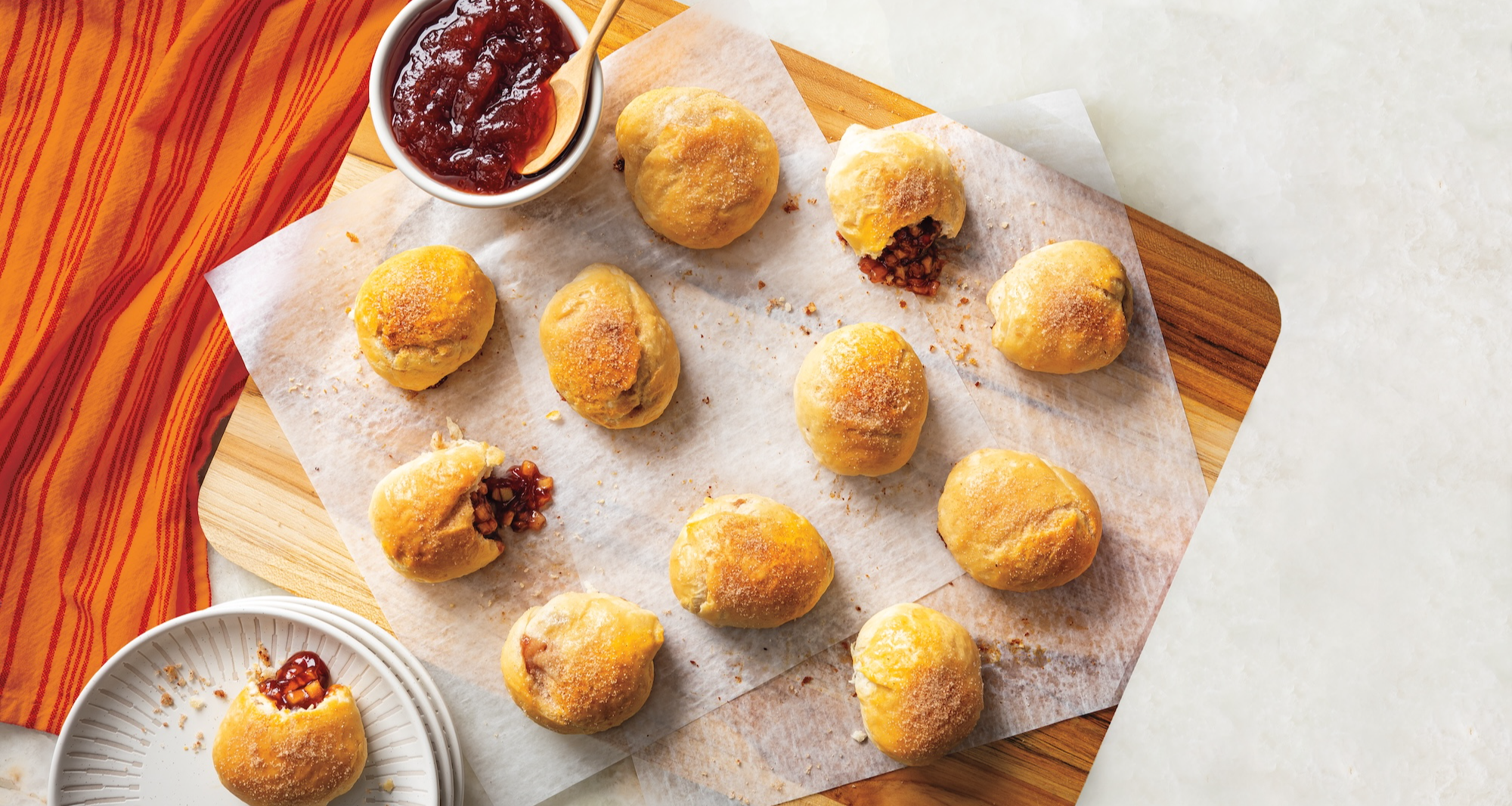 A cutting board holds several golden-brown mini biscuits on parchment paper, some split open to show the apple-cranberry filling. A bowl of cranberry sauce and a stack of small plates with one biscuit are also visible.