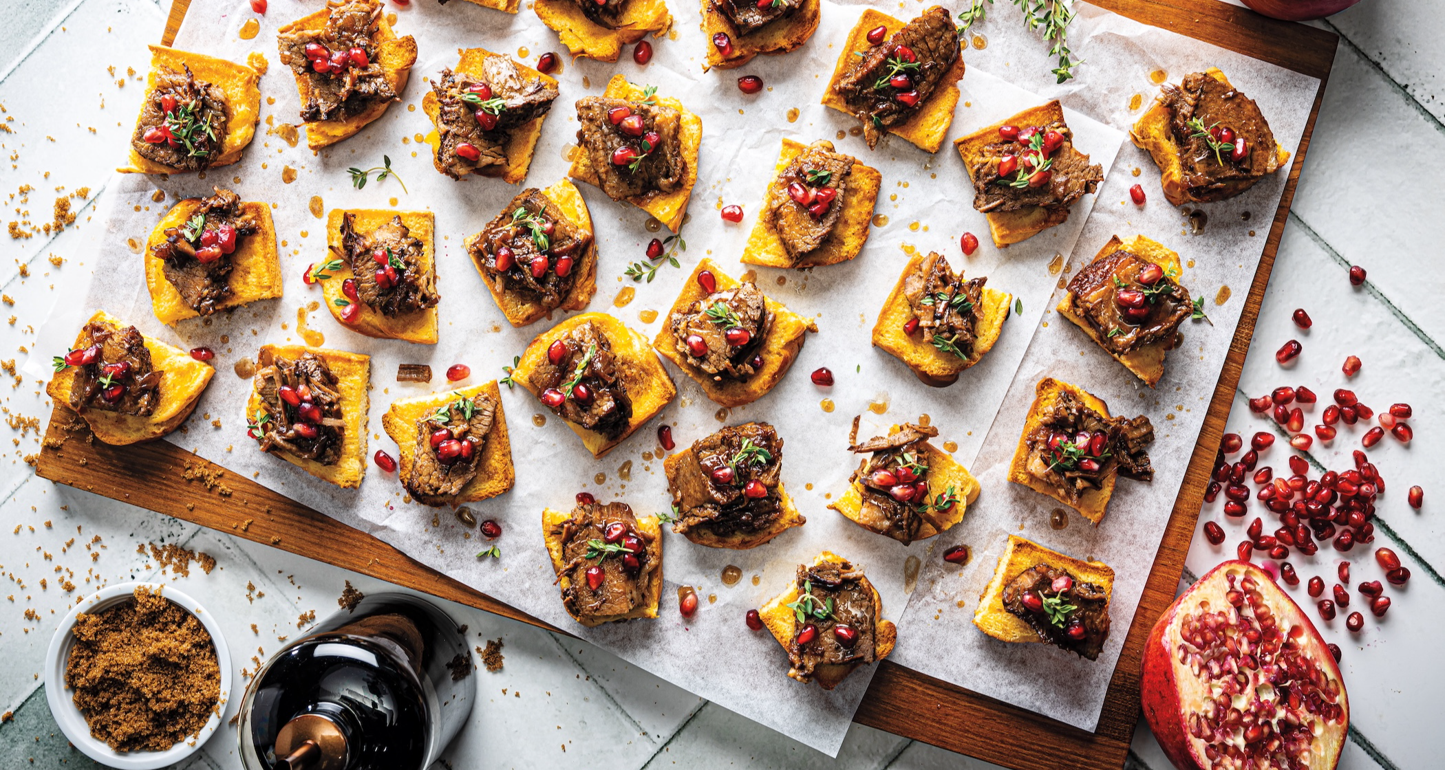 A wooden cutting board holds a sheet of parchment paper with several toasted challah bread squares topped with balsamic-glazed brisket, pomegranate arils, and thyme. A half pomegranate, an onion, and brown sugar are nearby.