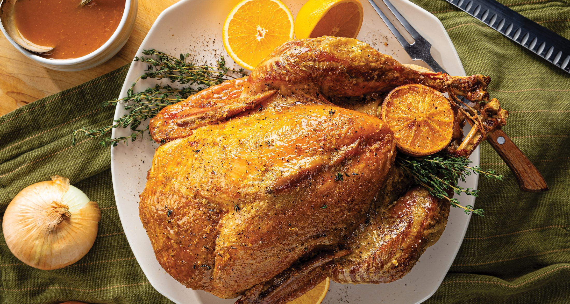 A beautifully roasted turkey, garnished with orange slices and thyme sprigs, is centered on a white platter. Two whole onions and a bowl of jus are placed on a green cloth and wooden board beside the platter.