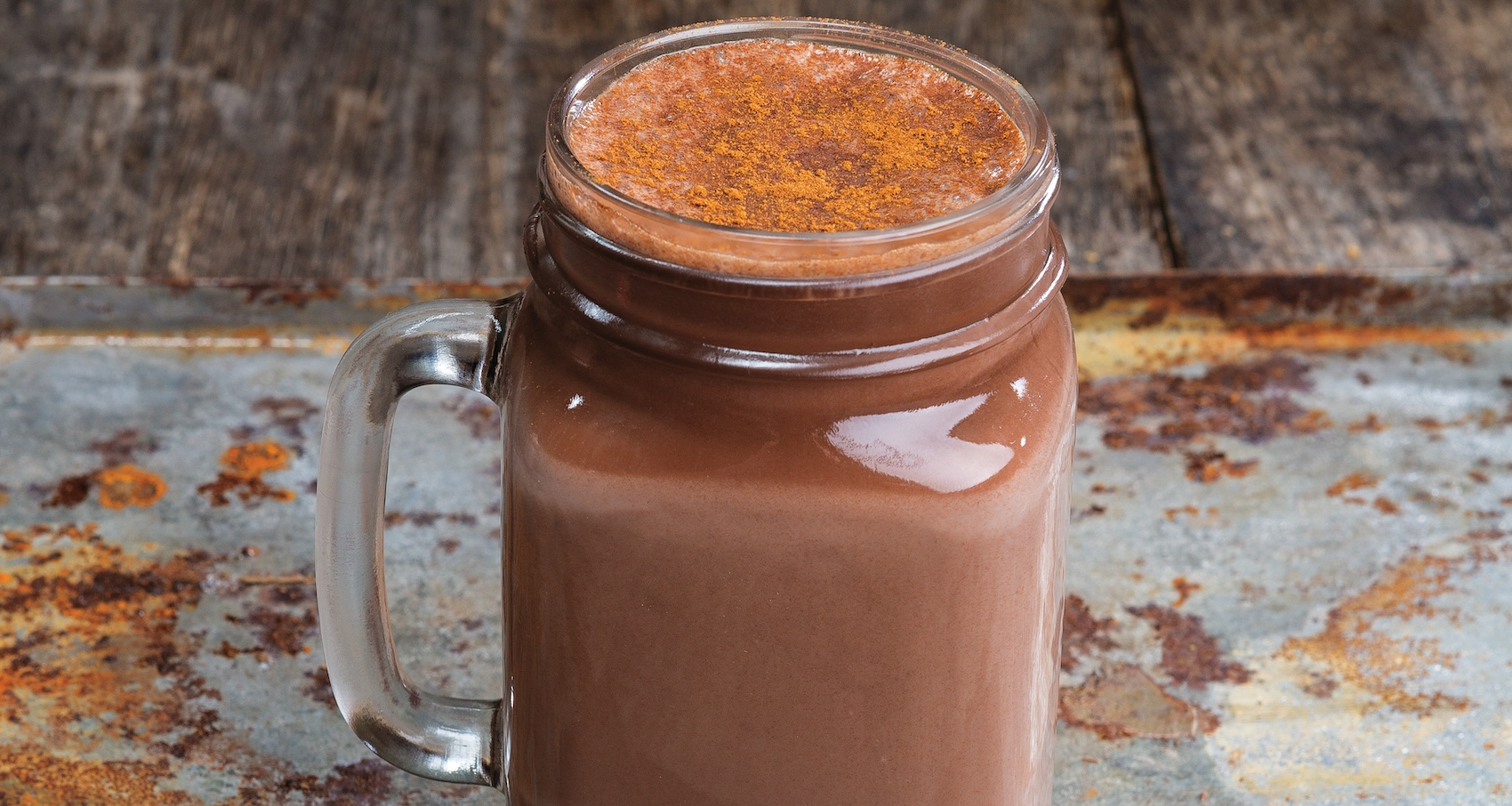 A glass mason jar mug of light brown Chai Hot Chocolate dusted with cinnamon is on a rustic metal tray. Two squares of dark chocolate, cardamom pods, a cinnamon stick, and peppercorns are scattered around the base.