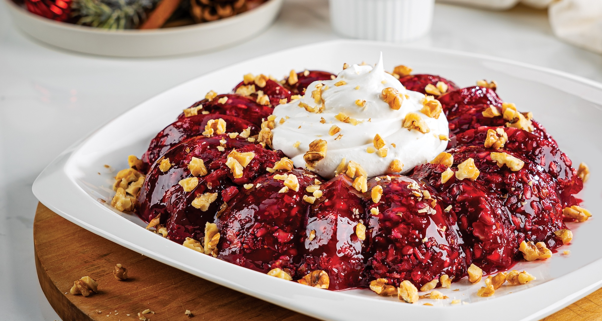 A dark red cranberry gelatin ring on a white plate is topped with a mound of whipped cream and chopped walnuts. Red Christmas ornaments, pine cones, and a glass of pomegranate seeds sit in the background