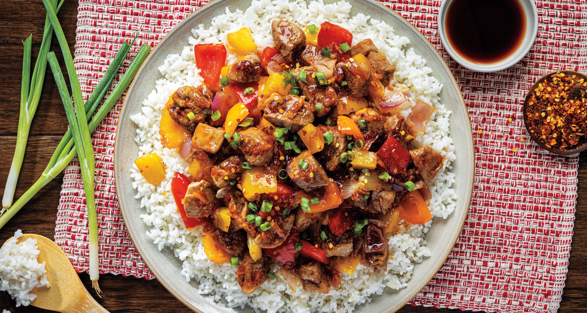A plate of white rice is topped with dark-glazed sweet and sour pork, red and yellow bell peppers, and fresh pineapple chunks. A small bowl of soy sauce, crushed red pepper flakes, and green onions are nearby.
