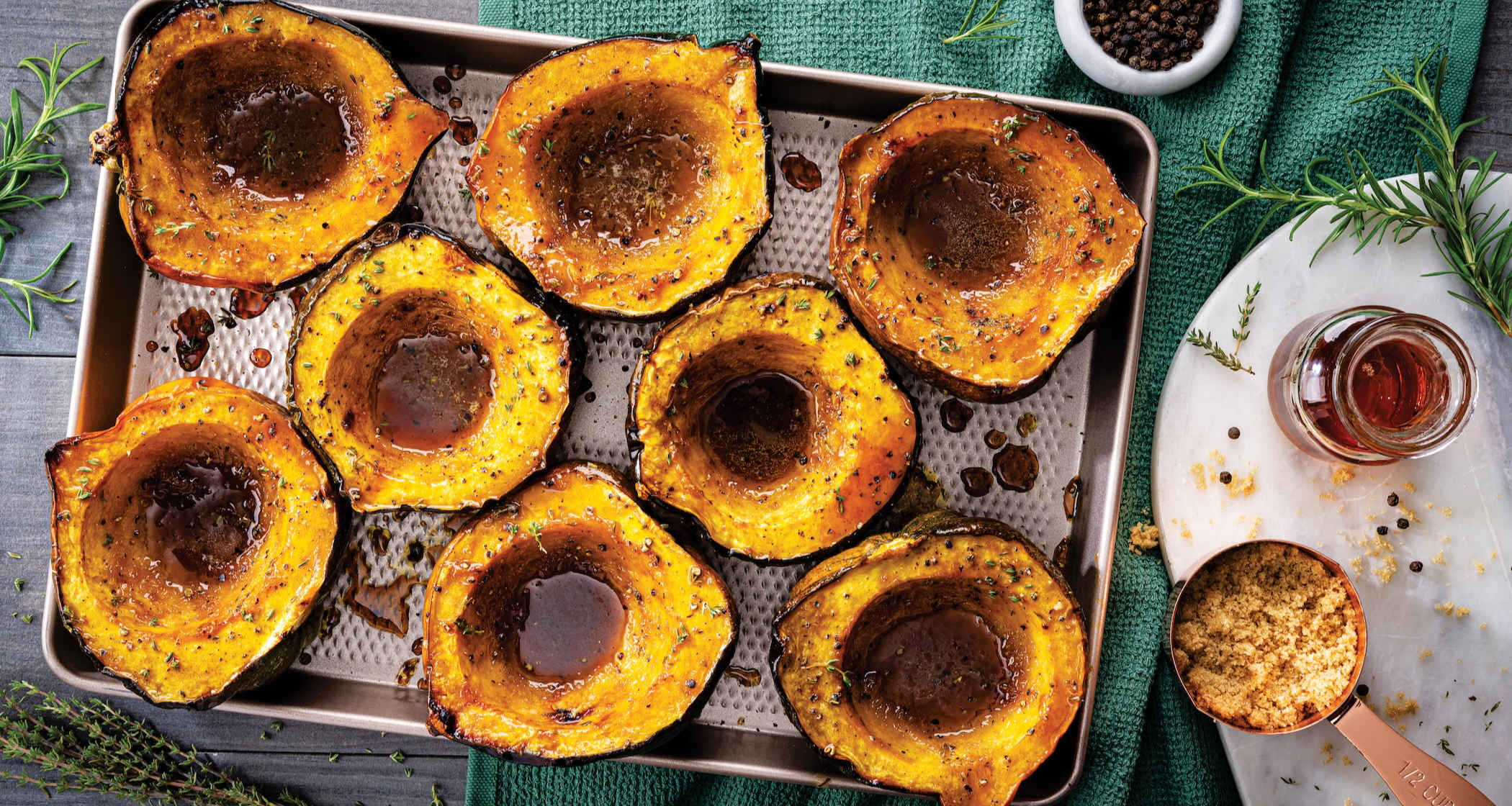 A metal baking sheet holds eight golden-yellow halves of acorn squash, roasted and filled with a brown maple glaze and herbs. Brown sugar, maple syrup, and rosemary are visible on a white marble board below.