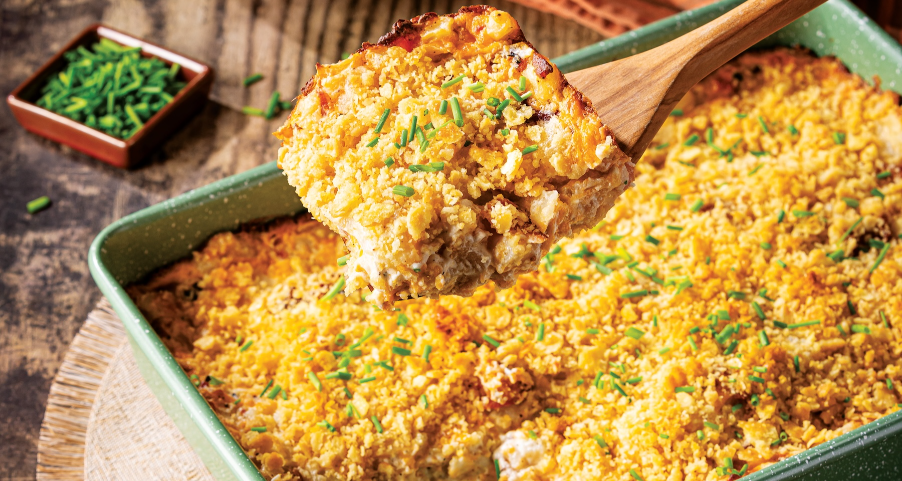 A square green baking dish holds a potato casserole with a golden-brown cracker topping sprinkled with chives. A wooden spoon lifts a serving out of the dish. Stacks of grey plates and a small pumpkin are in the background.