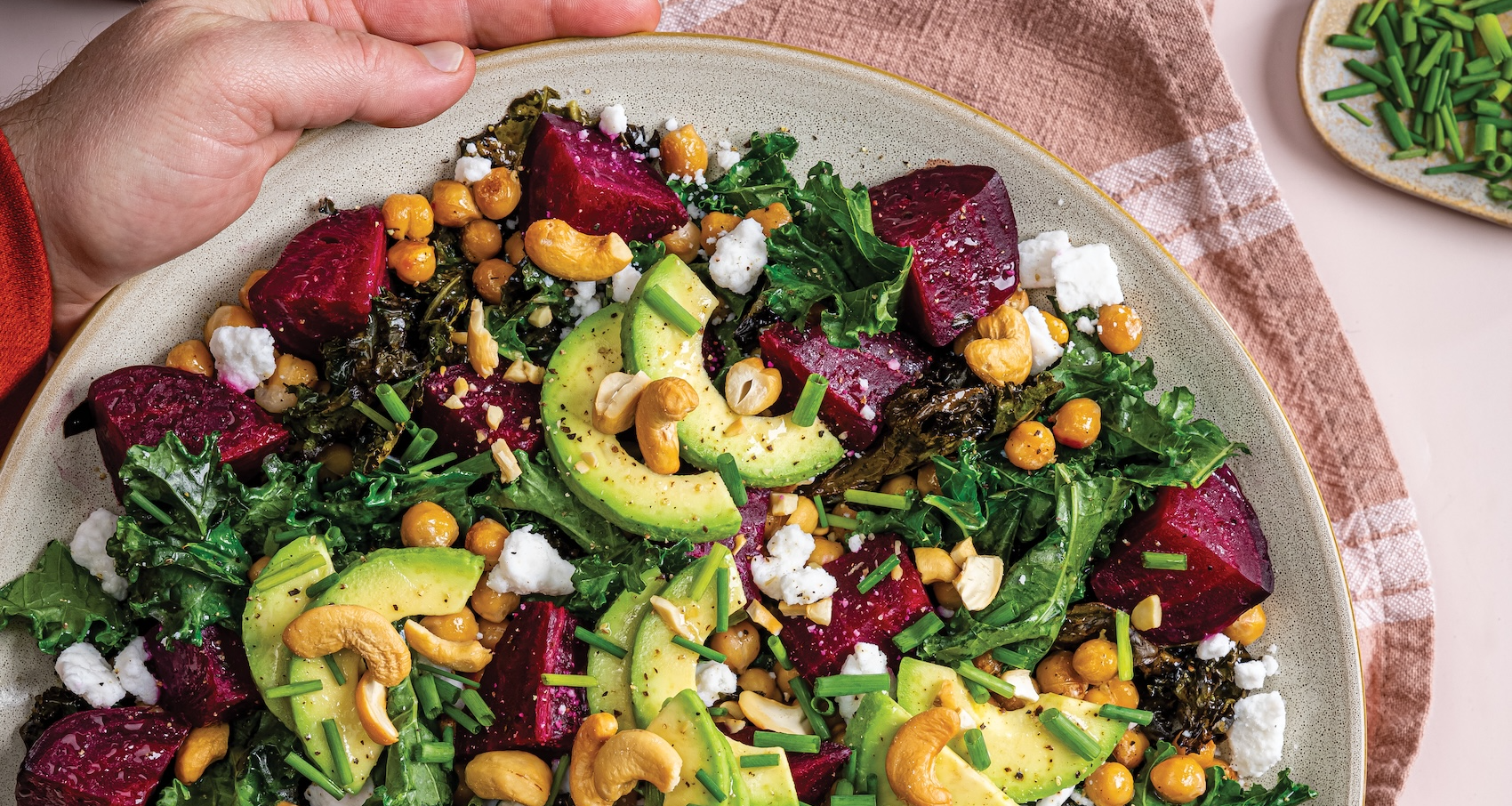 A large white plate holds a vibrant salad of dark green kale, red roasted beets, garbanzo beans, and creamy avocado slices. The salad is topped with white plant-based feta crumbles, chives, and whole salted cashews.