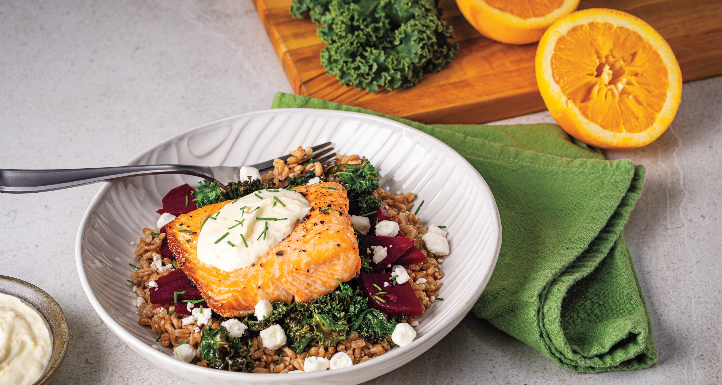 A white bowl holds a serving of savory salmon over farro, kale, and diced red beets. The salmon is topped with a white orange-yogurt sauce and chives. A fork, an orange half, and a green napkin are beside the bowl.