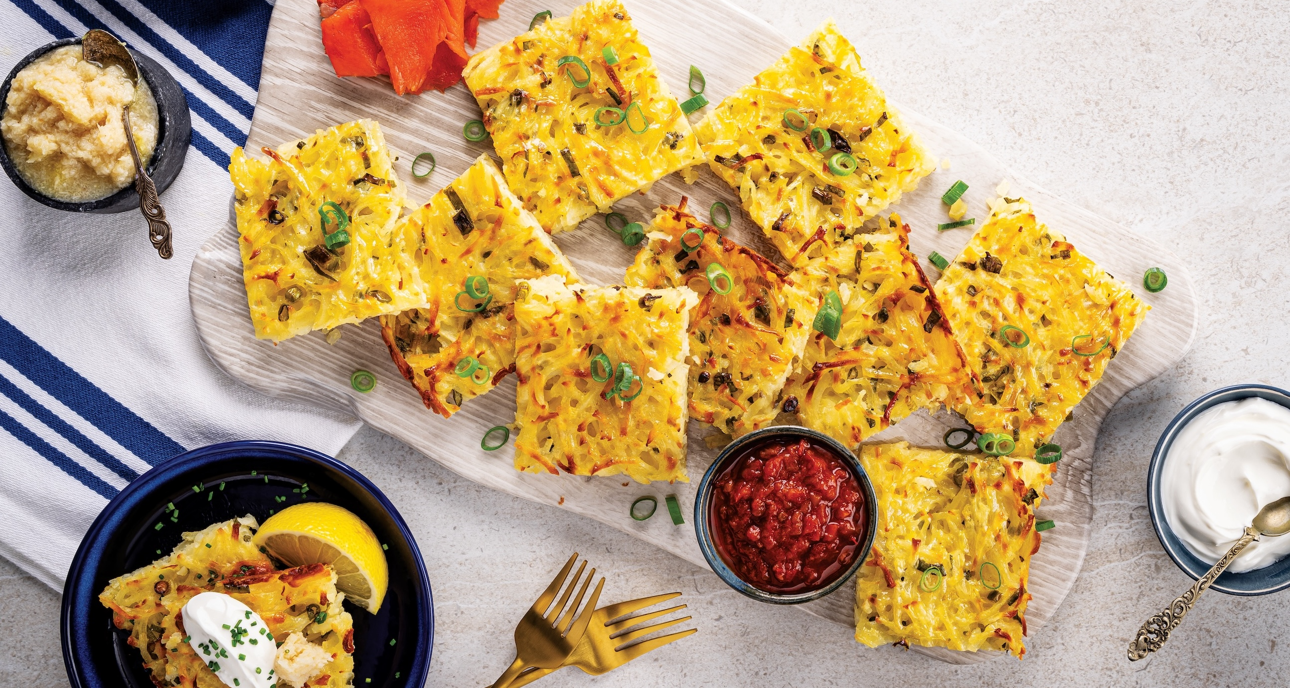 A cutting board holds squares of golden-brown latkes topped with green onions. Bowls of applesauce, horseradish, sour cream, and red relish surround the potatoes.