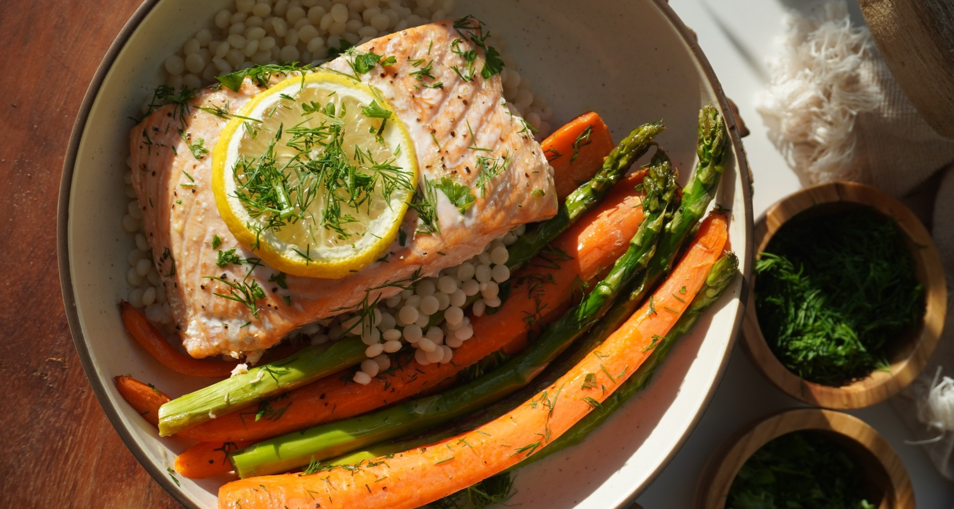 A bowl with baked salmon topped with lemon and fresh herbs, served over pearled couscous with roasted carrots and asparagus.