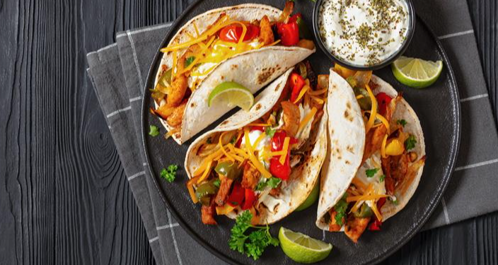 Three chicken fajitas in flour tortillas topped with melted cheddar cheese, sautéed peppers, and onions, served on a black plate with lime wedges and a side of sour cream.