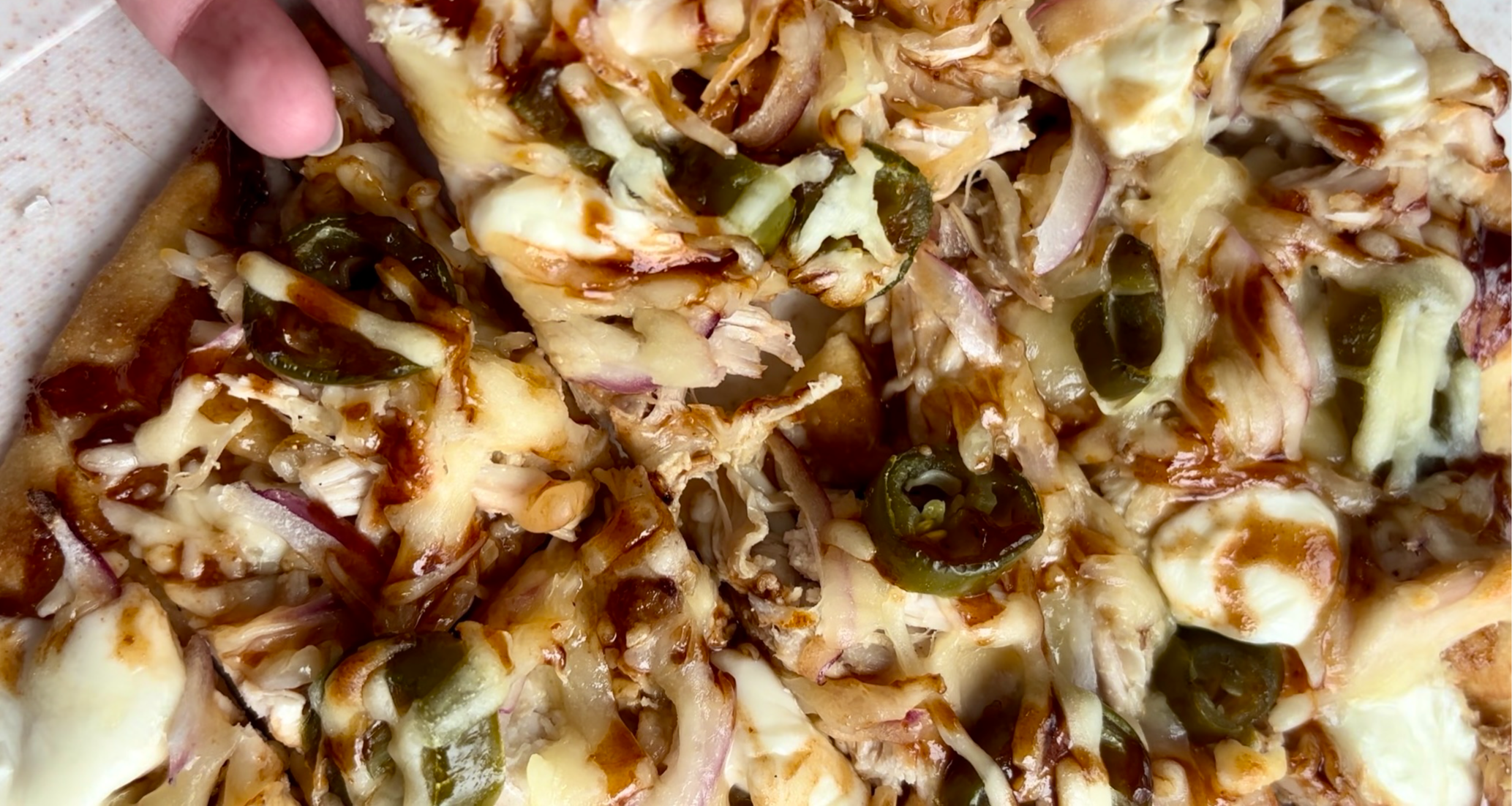 Close-up of a crispy BBQ rotisserie chicken flatbread pizza topped with melted buffalo mozzarella, shredded cheese, thinly sliced red onions, and pickled jalapeños, finished with a tangy BBQ sauce drizzle.
