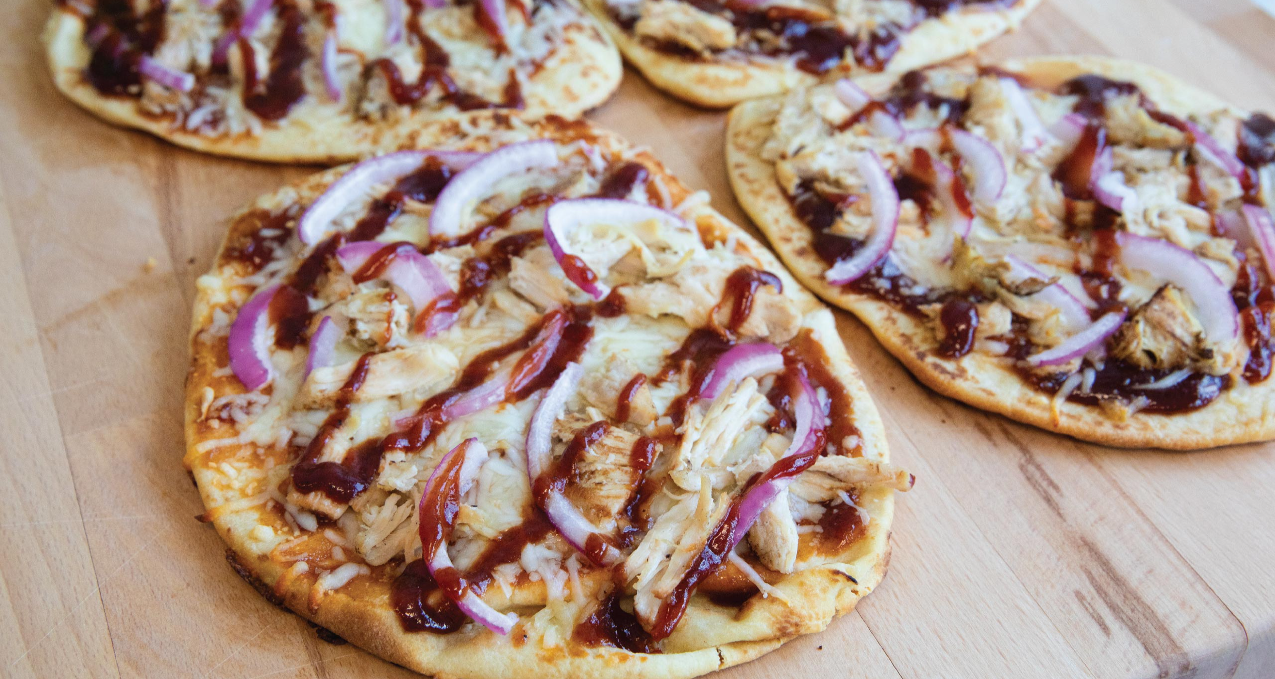 BBQ chicken flatbreads topped with melted cheese, red onion slices, and drizzled barbecue sauce, arranged on a wooden board.