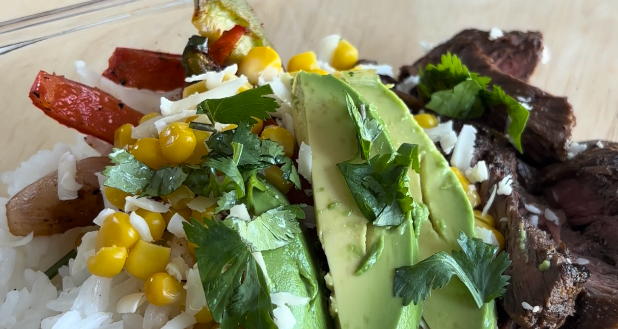 A top-down, close-up shot of a glass meal prep container filled with cilantro lime rice, roasted red peppers, steak strips, corn, and shredded cheese. Fresh avocado slices are being garnished with chopped cilantro.