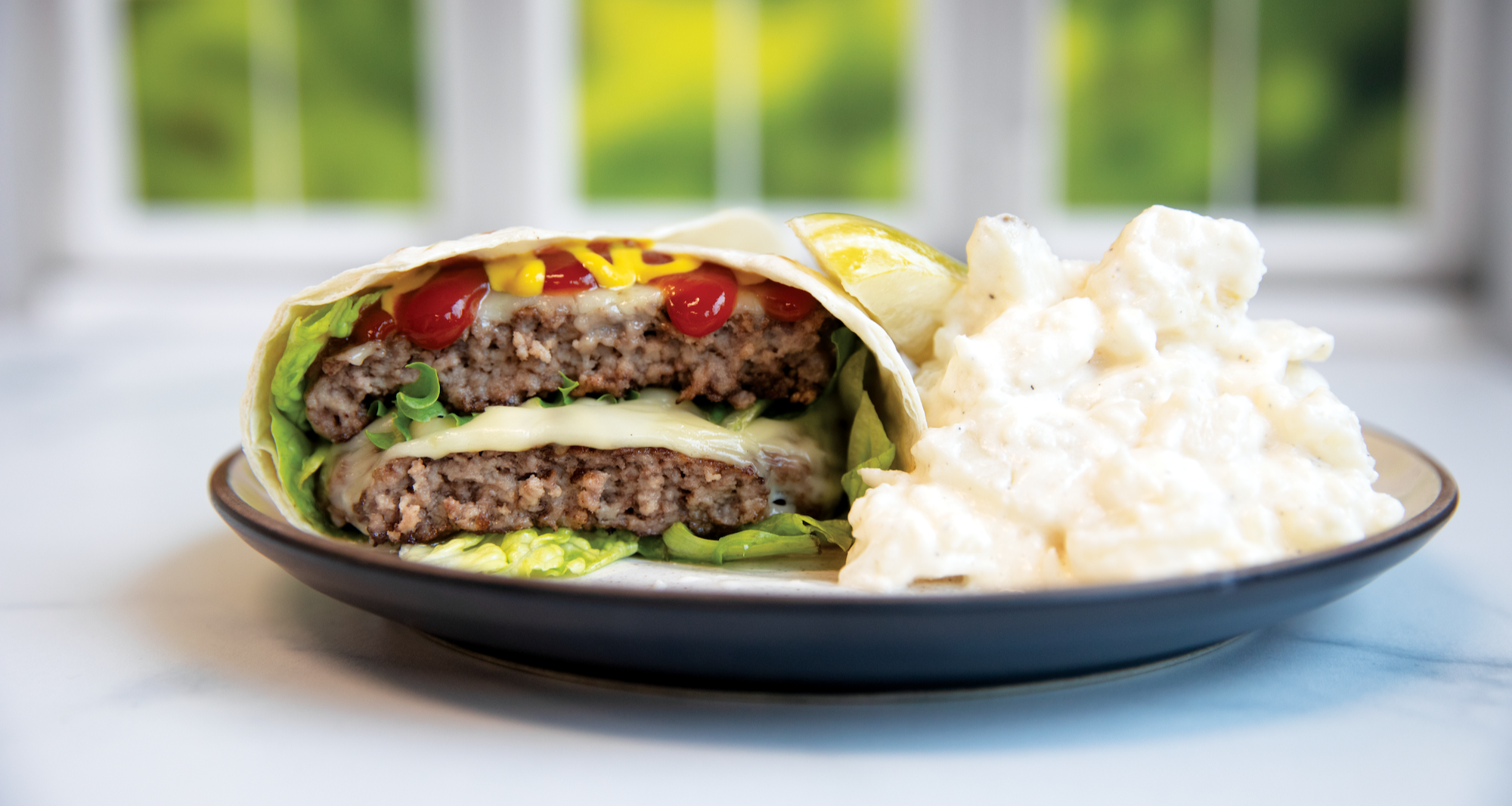Smashed cheeseburger wrap with lettuce, melted cheese, ketchup, and mustard, served with a side of potato salad on a plate.